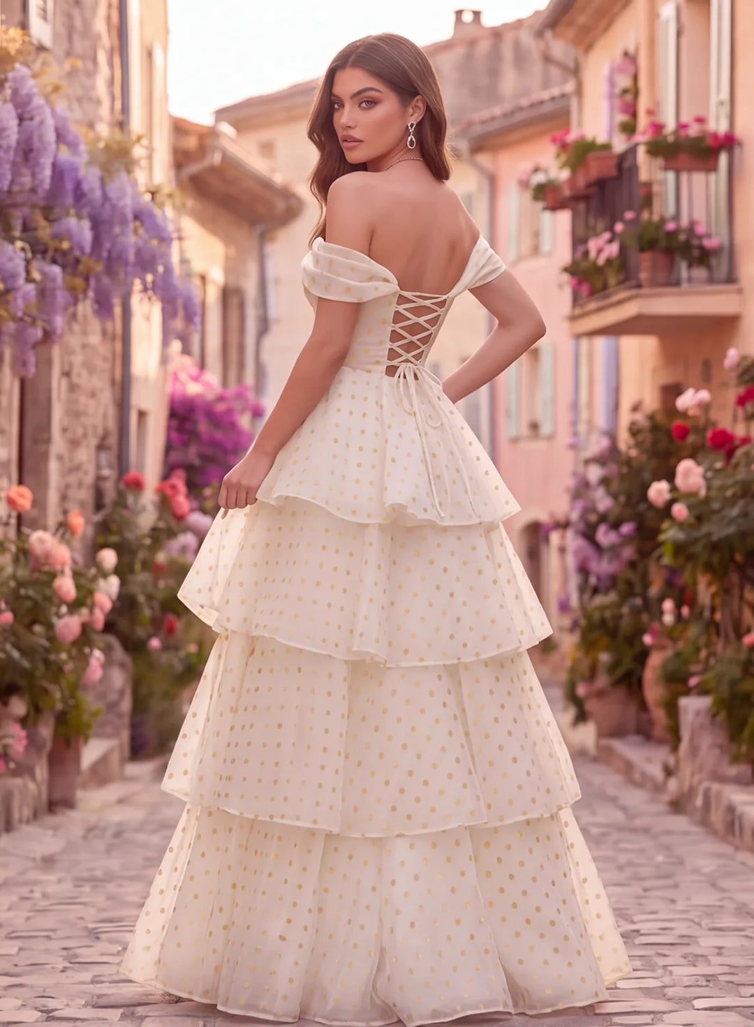 Back view of an ivory and gold polka dot off shoulder prom dress with a lace-up corset back and layered floor length skirt.
