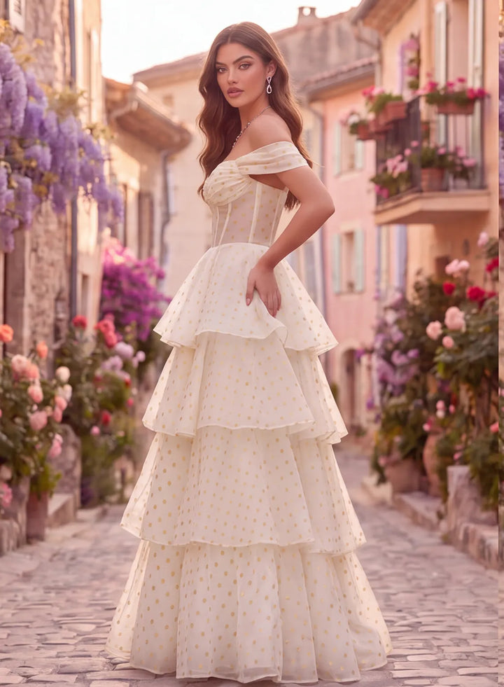 Side view of an ivory and gold polka dot prom gown featuring an off shoulder neckline, fitted corset bodice, and flowing tiered skirt.